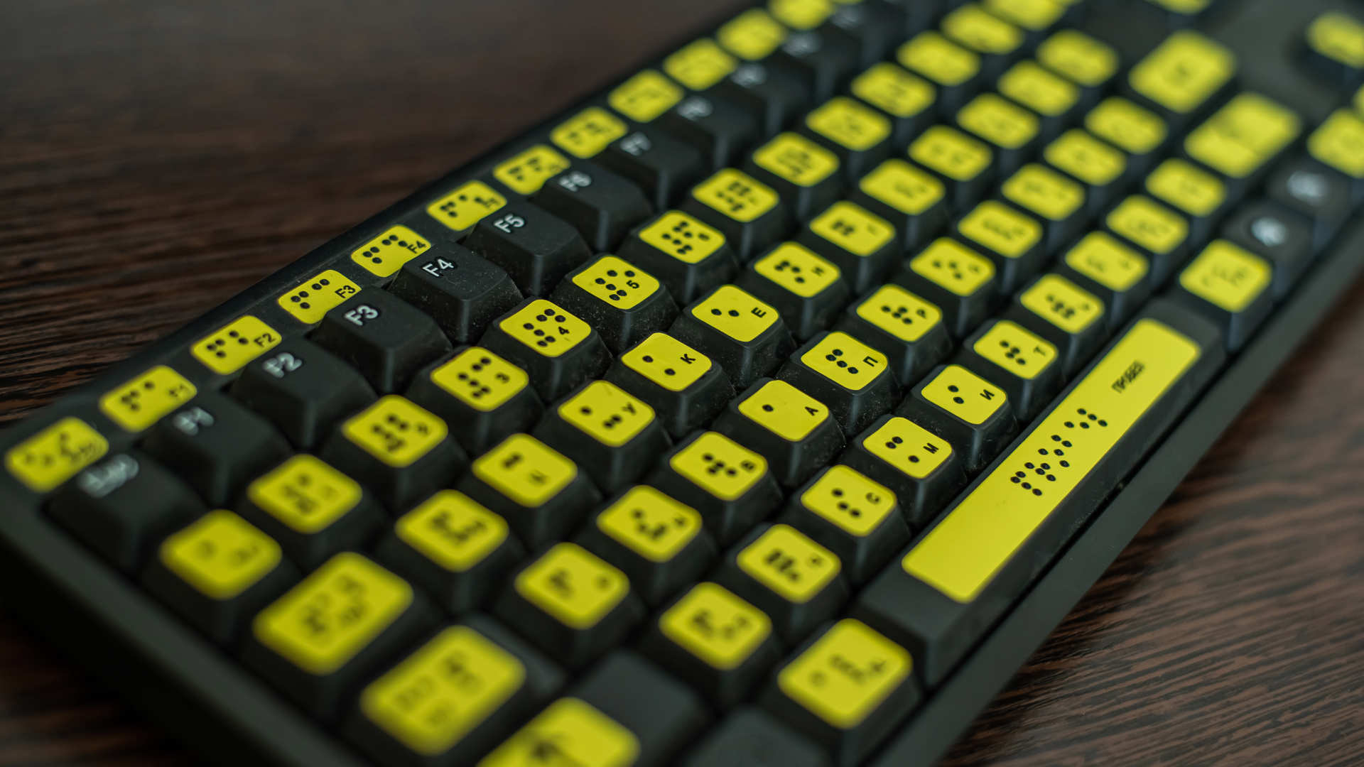 A black computer keyboard on a dark wooden desk, with vivid a yellow sticker on each key showing both the letter and Braille equivalent.