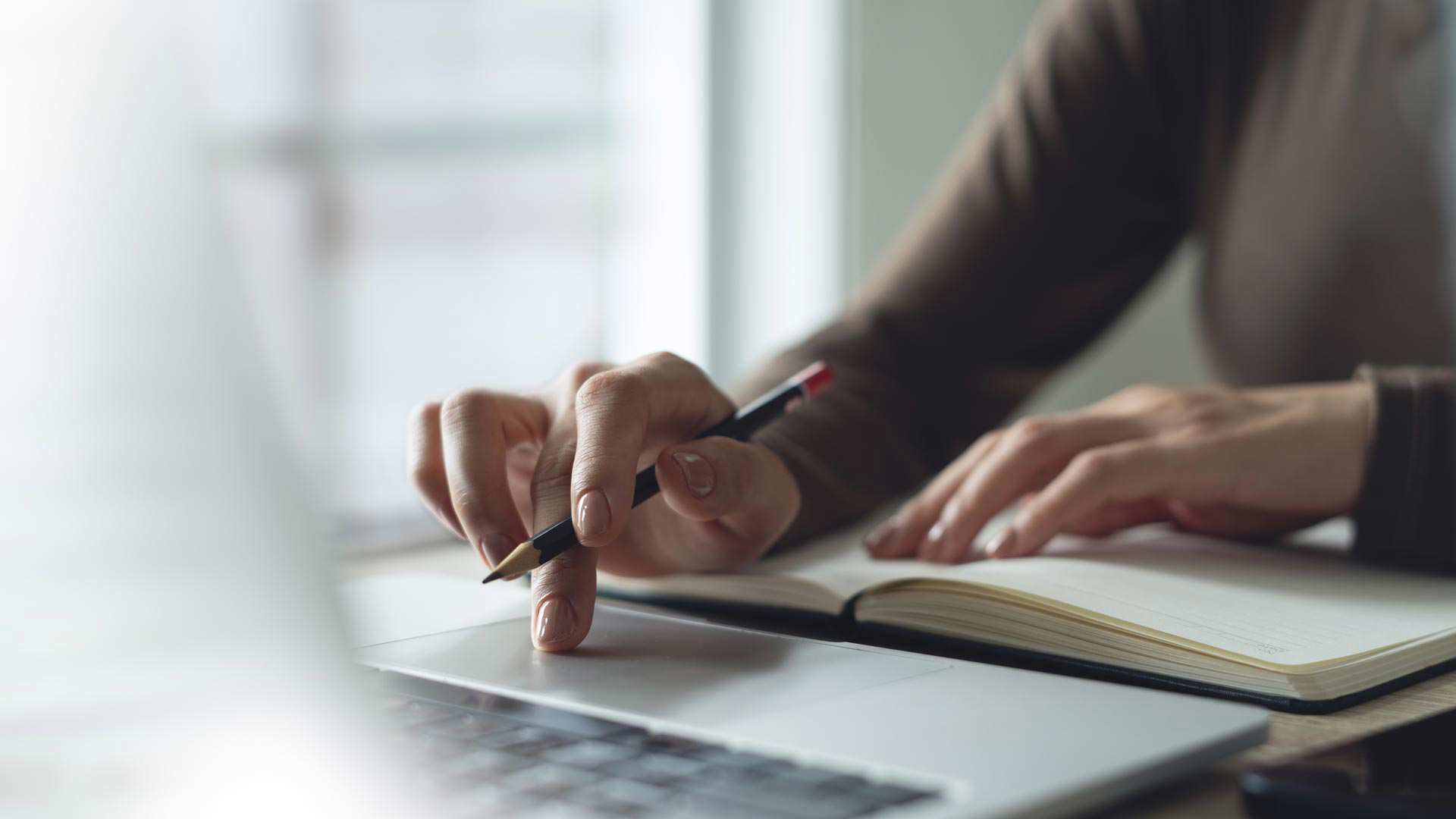 Hand of the person is pressing the keypad of the laptop, searching of the information with an open book.